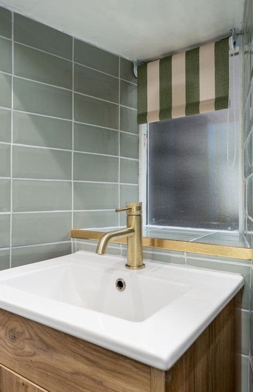 Modern bathroom with white sink, brass faucet, green tiled shower wall and striped window blind