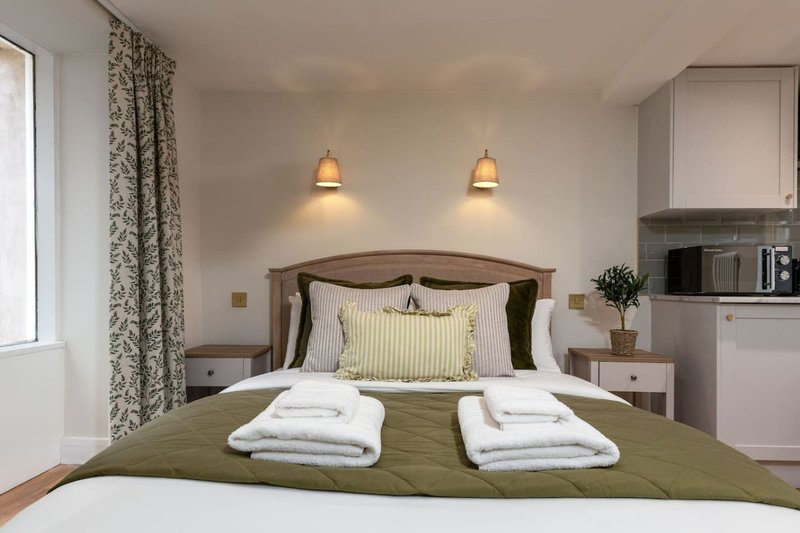 Modern bedroom with double bed, olive green bedding, pendant lights, patterned curtains and kitchenette