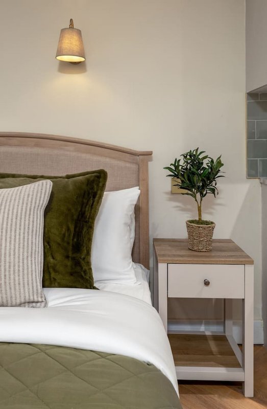 Bedroom with wooden bed frame, green cushions, white bedding, pendant lamp and potted plant on nightstand