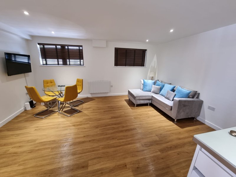 Spacious open-plan living area with wooden flooring, grey sofa with blue cushions, yellow armchairs, and wall-mounted TV