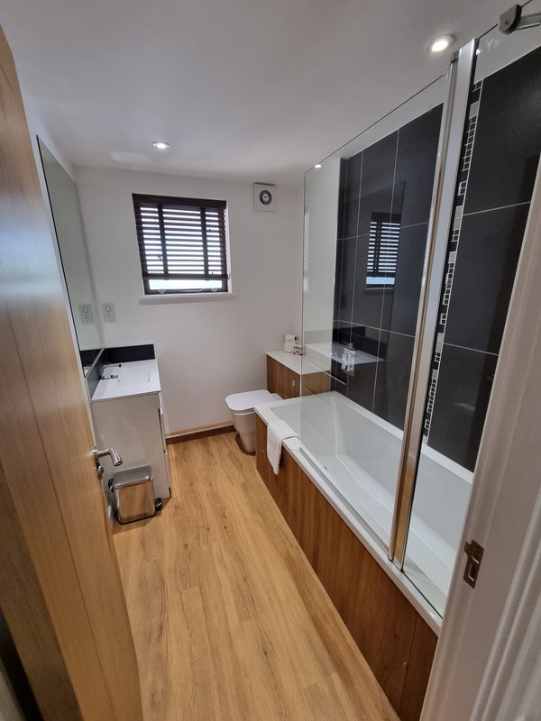 Modern bathroom with white bathtub, black tiled shower enclosure, wooden flooring and window