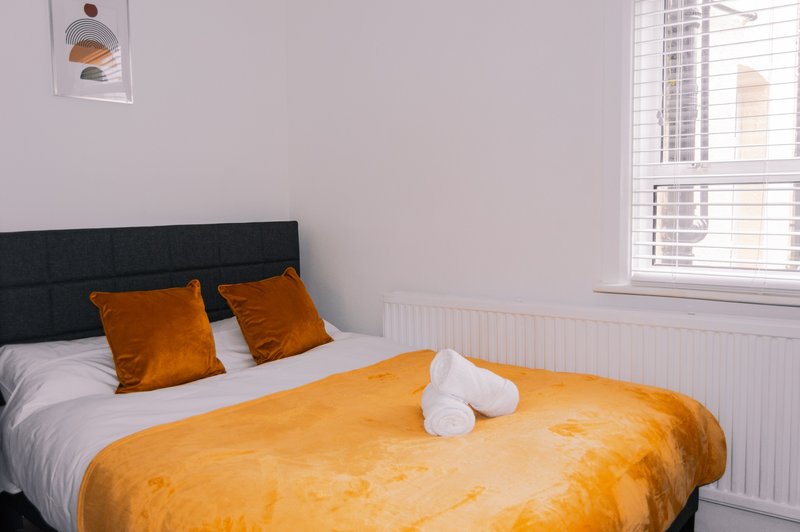 Bedroom with double bed, black headboard, orange bedding and cushions, window with blinds
