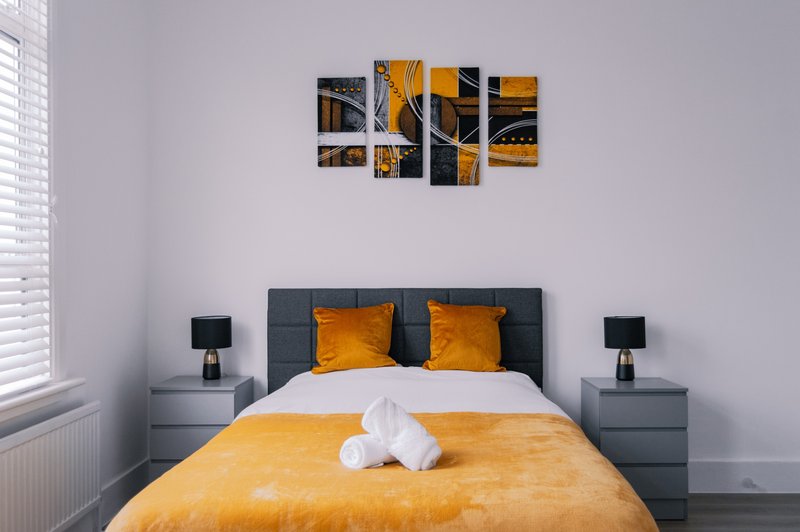 Modern bedroom with yellow and grey bedding, dark headboard, bedside tables and wall art