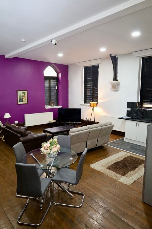 Modern living room with purple accent wall, gray sofa, glass coffee table, and kitchenette area