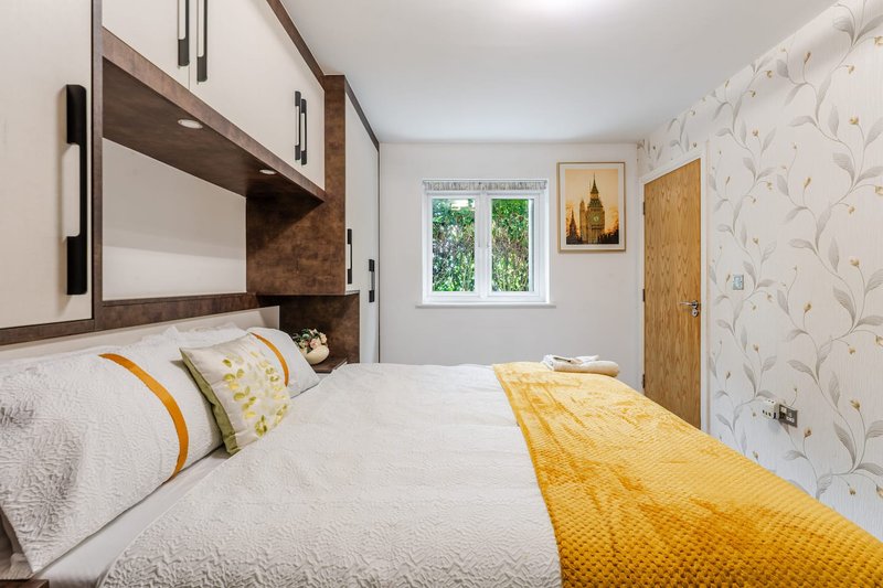 Bright bedroom with double bed, yellow throw, patterned wallpaper and wooden wardrobe