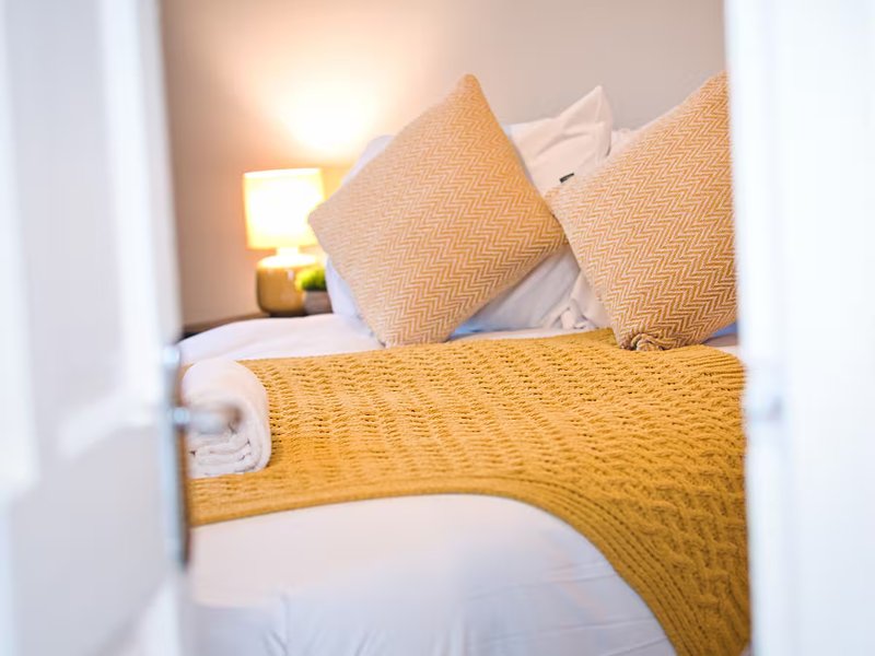Bedroom with double bed, yellow throw blanket, neutral cushions and bedside lamp