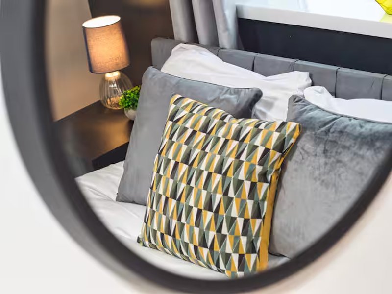 Modern bedroom with gray upholstered headboard, patterned yellow and gray cushions, bedside lamp and white linens