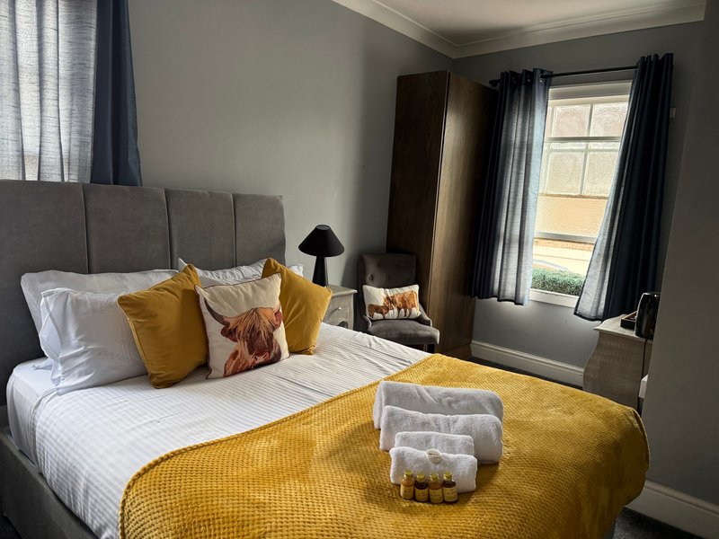 Bedroom with grey upholstered headboard, double bed, yellow bedding, cushions and window with curtains