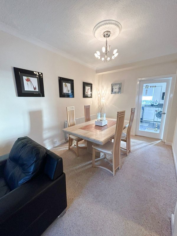 Open-plan dining and living area with wooden table, chairs, black sofa, framed wall art, and glass door to kitchen