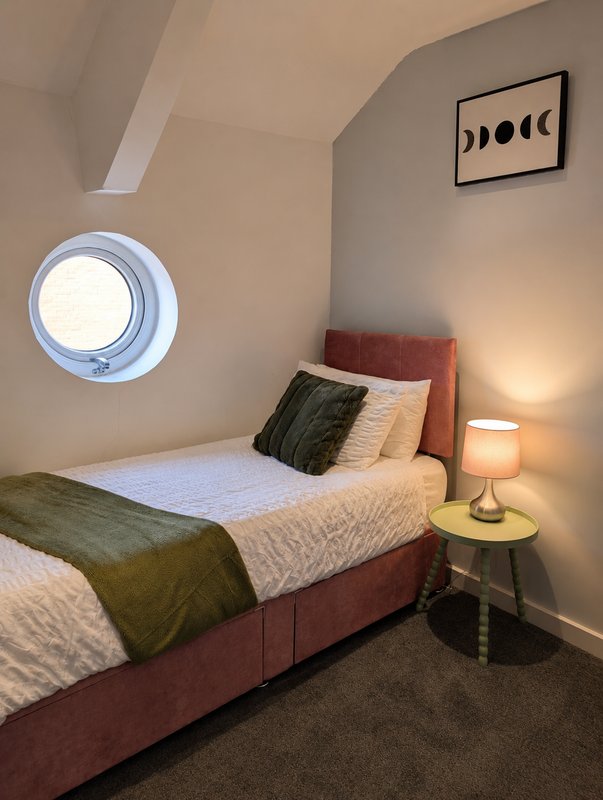 Bedroom with single bed, burgundy headboard, round window, bedside lamp and wall art
