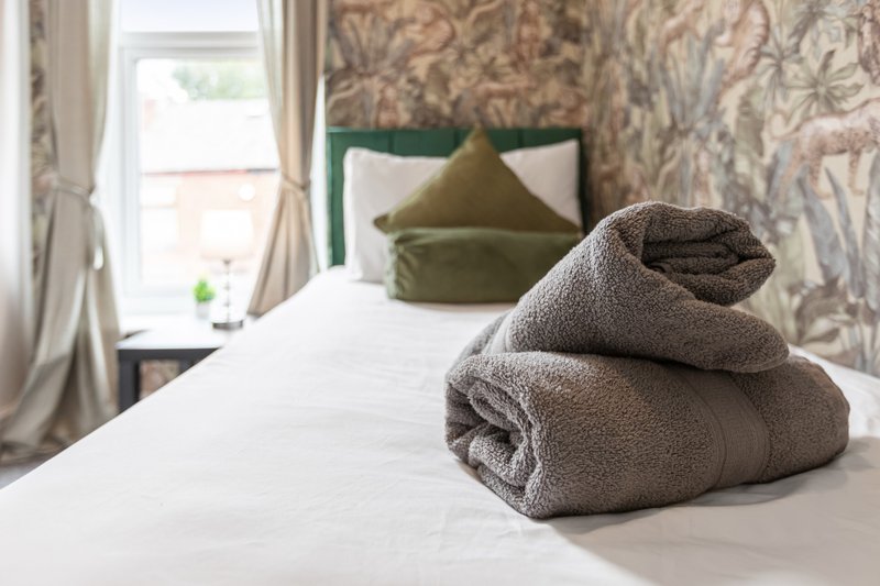 Bedroom with neatly folded towels on bed, green headboard, patterned wallpaper and window