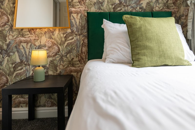 Bedroom with green upholstered headboard, white bedding, patterned wallpaper and black nightstand