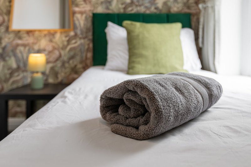 Bedroom with rolled gray towel on bed, green headboard, white pillows, and patterned wallpaper