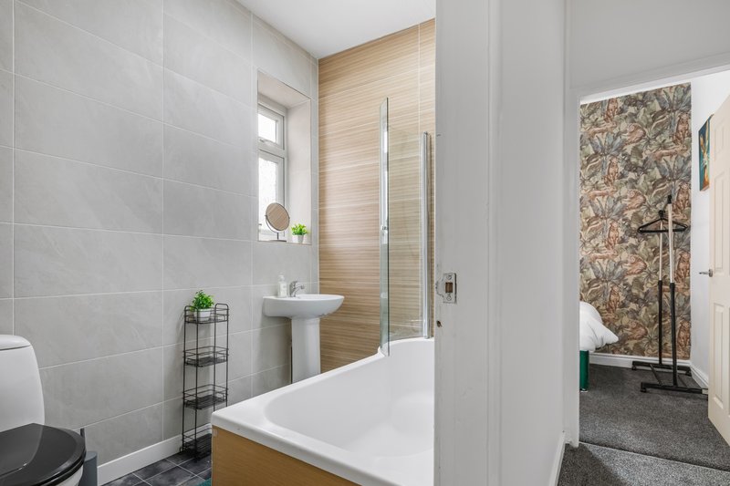 Modern bathroom with white bathtub, pedestal sink, grey tiles and wooden accents