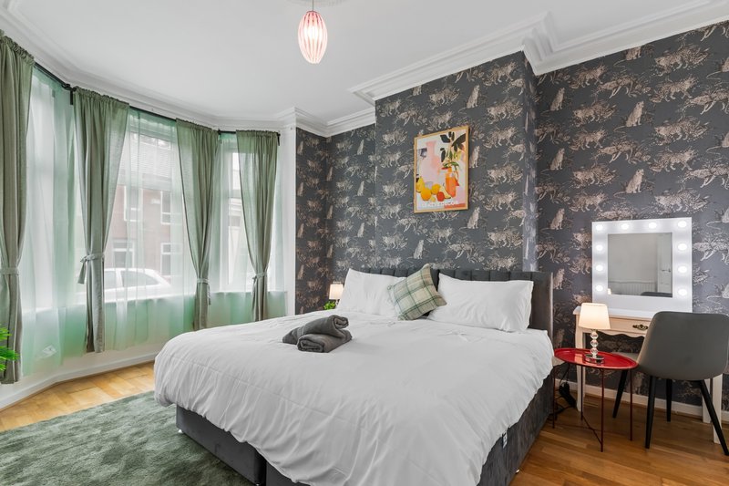 Bedroom with double bed, dark patterned wallpaper, green curtains, wooden floor and desk area