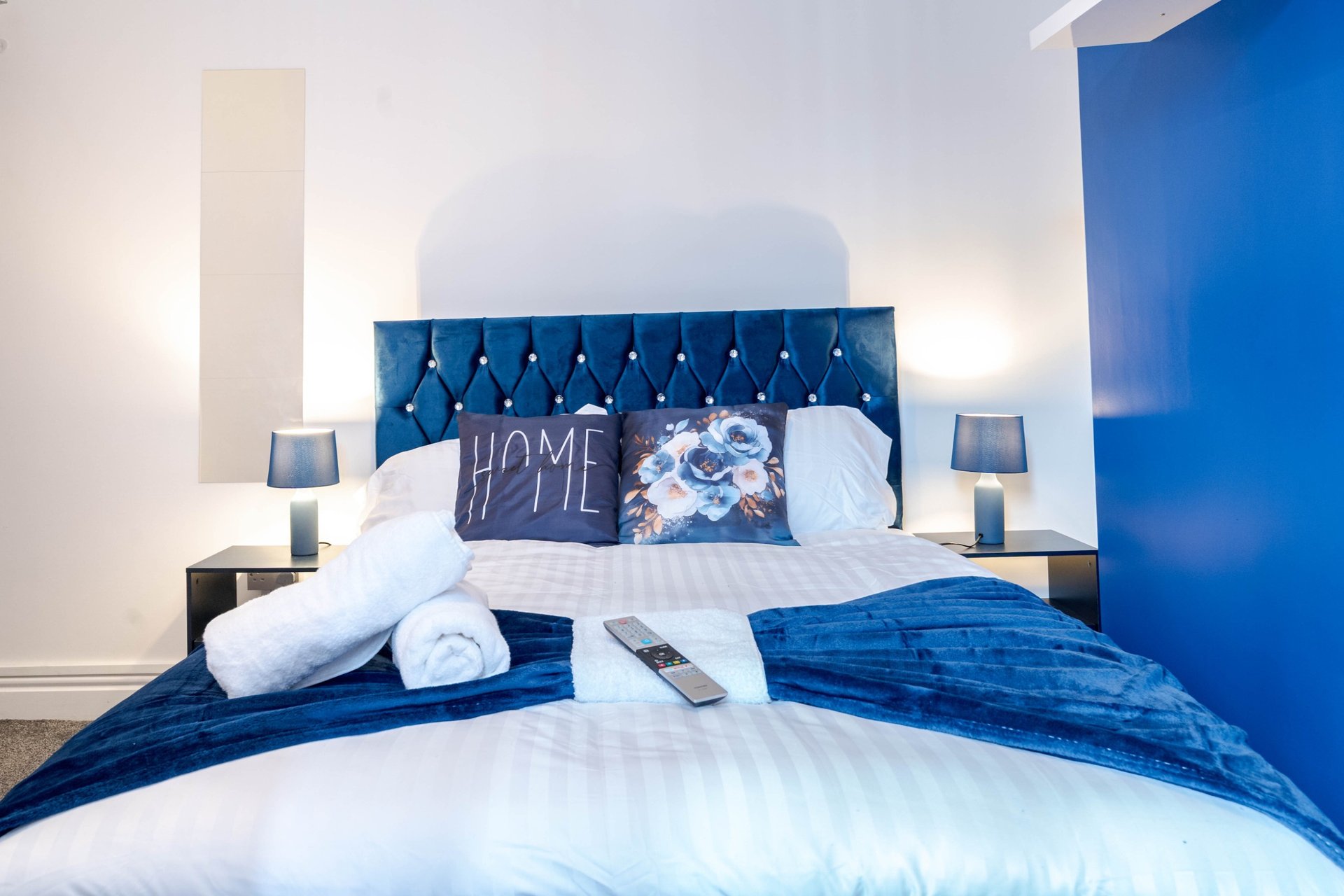 Modern bedroom with blue tufted headboard, white bedding, rolled towels, and decorative pillows