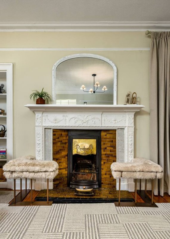 Living room with fireplace, white marble mantelpiece, arched mirror, and two upholstered stools