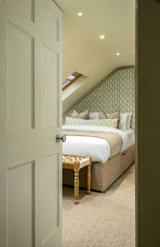 Bedroom with double bed, patterned headboard, white bedding, and gold footstool visible through open doorway
