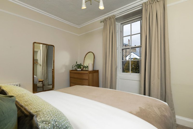 Bright bedroom with double bed, wooden dresser, mirrors, and large window with curtains