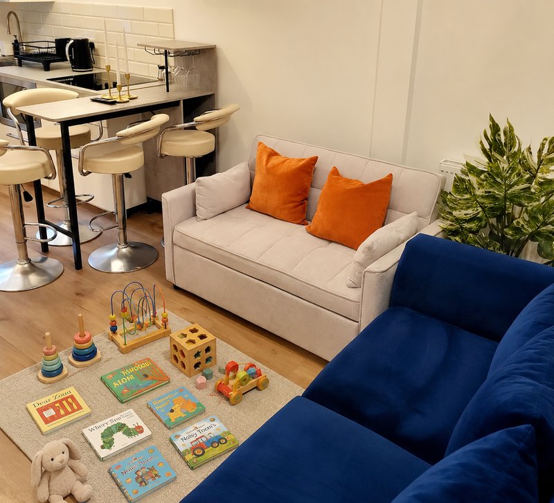 Open-plan living area with cream sofa, blue armchairs, kitchen counter, wooden flooring and toys