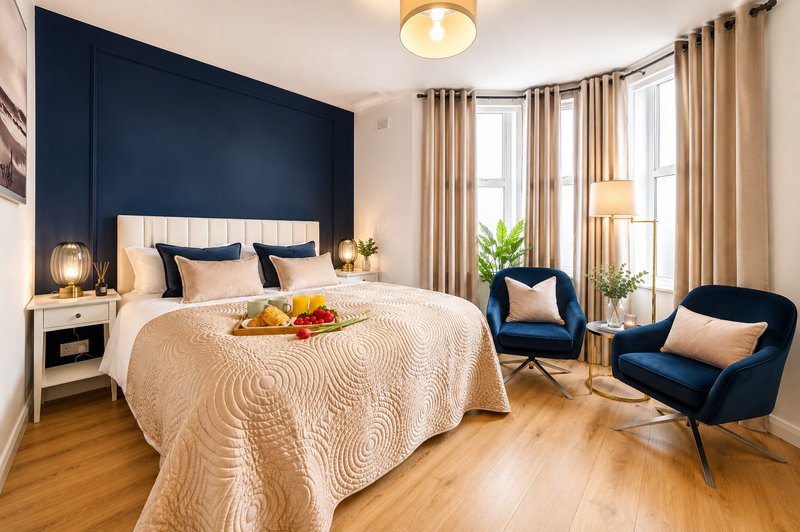 Modern bedroom with navy accent wall, double bed, two blue armchairs, wooden flooring and large windows