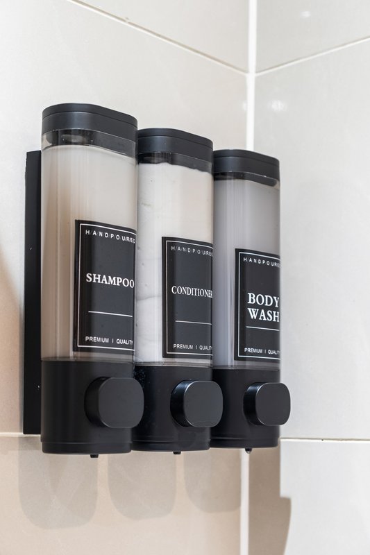 Wall-mounted black dispenser with three labeled bottles of shampoo, conditioner, and body wash