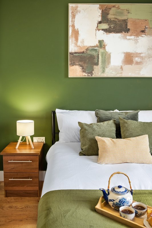 Bedroom with green walls, wooden nightstand, bed with green and cream pillows, framed abstract art