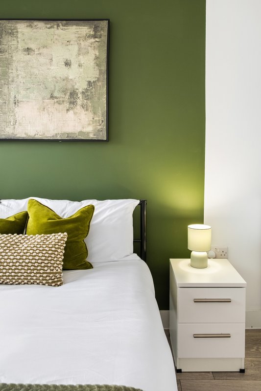 Bedroom with green accent wall, white bed, olive cushions, and bedside lamp
