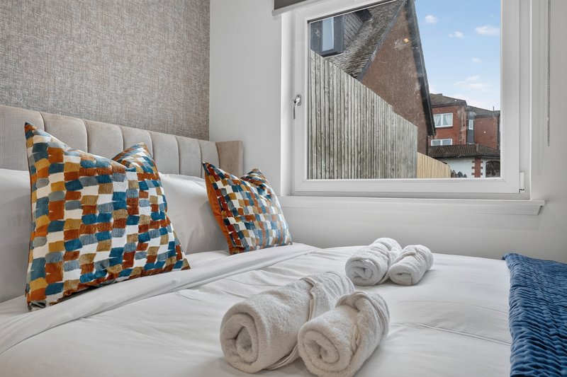 Bedroom with grey headboard, patterned cushions, rolled towels on white bedding, and window view of brick buildings