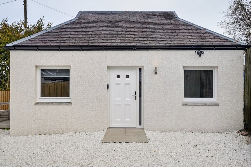Modern single-story cottage with cream render, dark roof, white door and two windows, gravel forecourt.