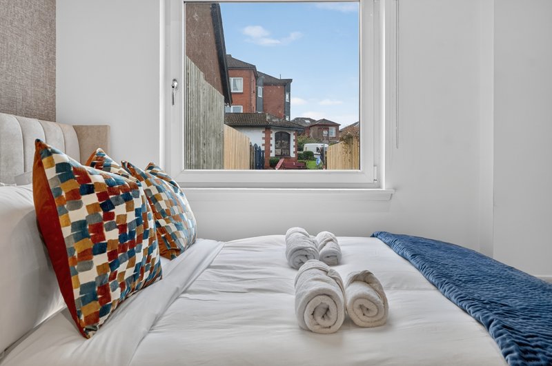 Bright bedroom with double bed, colorful cushions, rolled towels, and window overlooking brick buildings