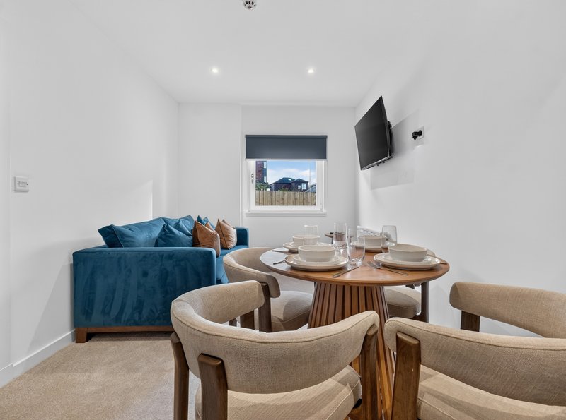 Open-plan living and dining area with blue sofa, wooden dining table, beige chairs and wall-mounted TV