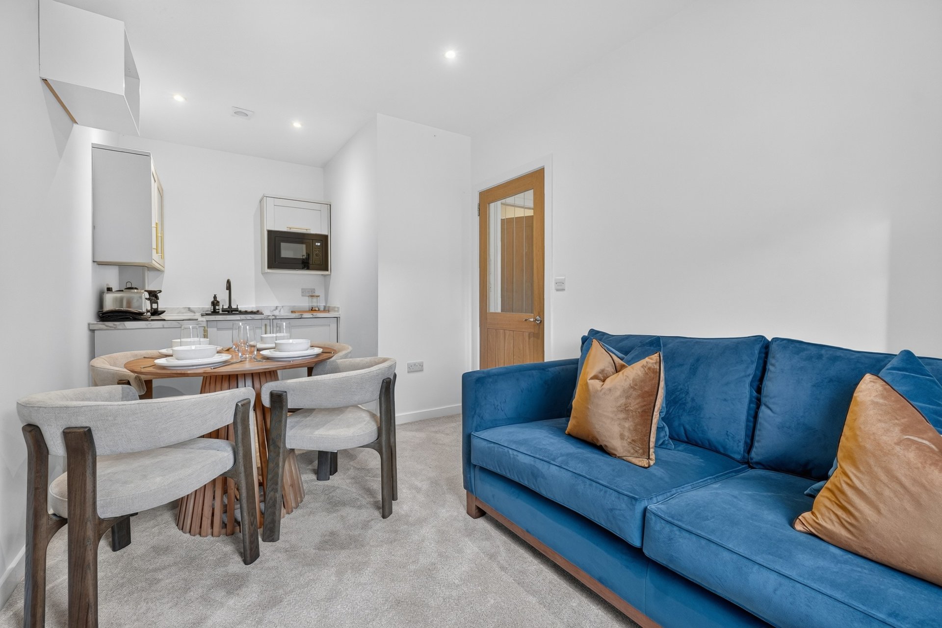 Open-plan living area with blue sofa, dining table, chairs, and kitchenette with wooden door