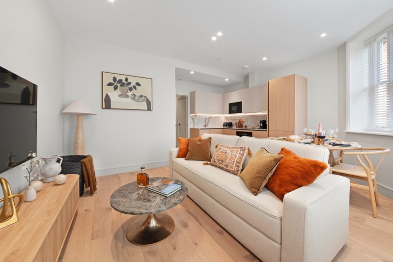 Open-plan living room with cream sofa, orange cushions, wooden furniture and kitchen visible