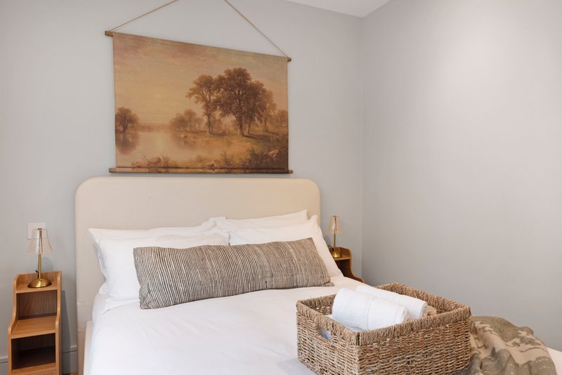 Bedroom with cream upholstered bed, white bedding, woven basket, and landscape artwork on wall