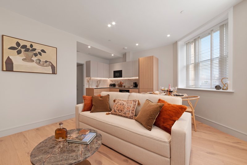 Open-plan living room with cream sofa, orange cushions, wooden flooring, and kitchenette