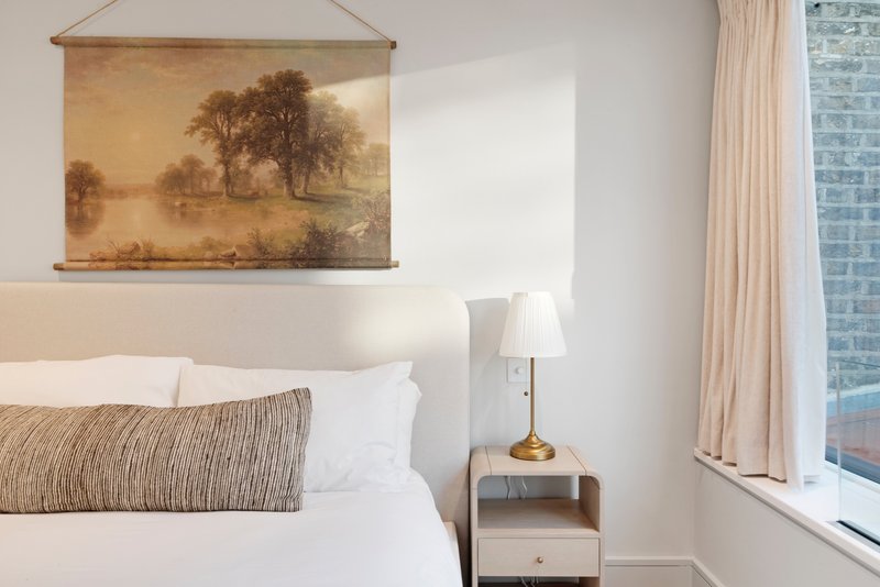Bedroom with double bed, white pillows, striped cushion, bedside table with lamp and framed landscape artwork on wall