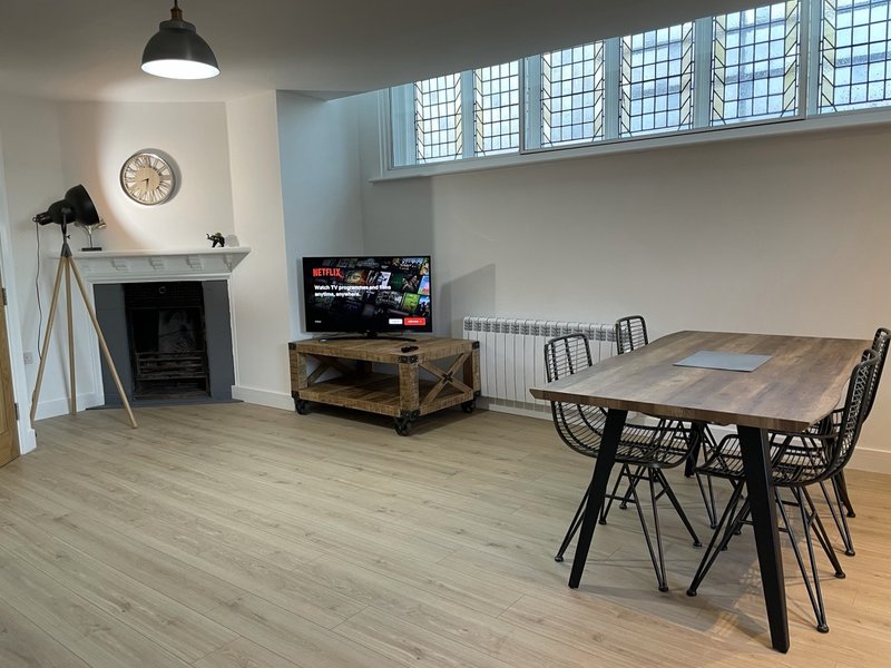 Open-plan living space with dining table, fireplace, TV, and high windows with natural light