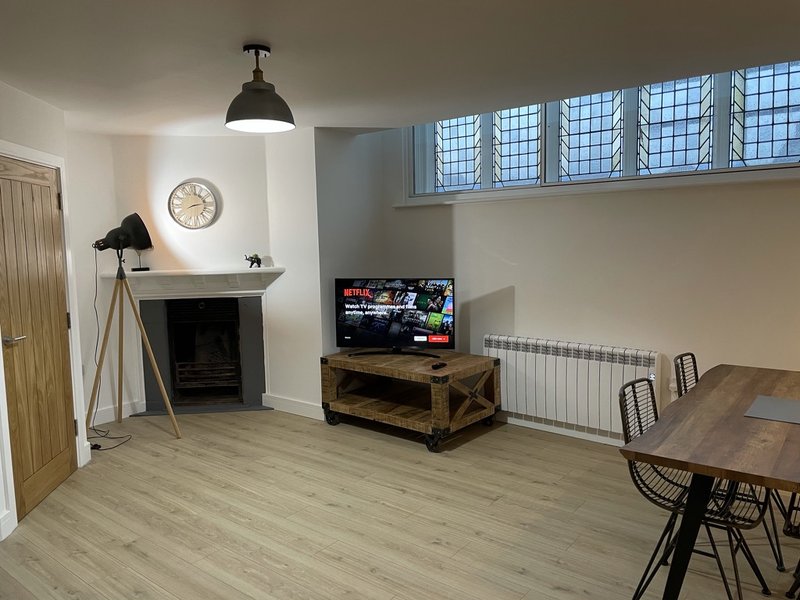 Open-plan living room with fireplace, TV on wooden stand, dining table and high windows