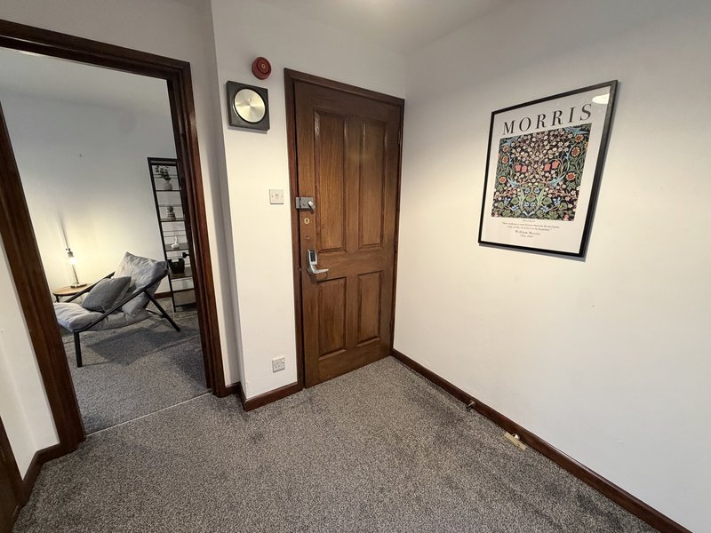 Hallway with wooden door, wall clock, framed artwork, and doorway to bedroom