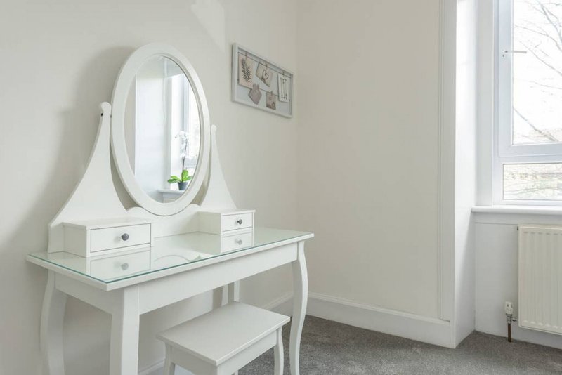 Bright bedroom corner with white dressing table, oval mirror, and matching stool
