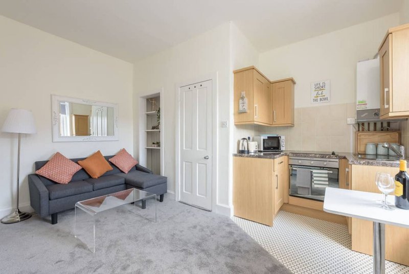 Open-plan living space with grey sofa, kitchen area with wooden cabinets and stainless steel appliances