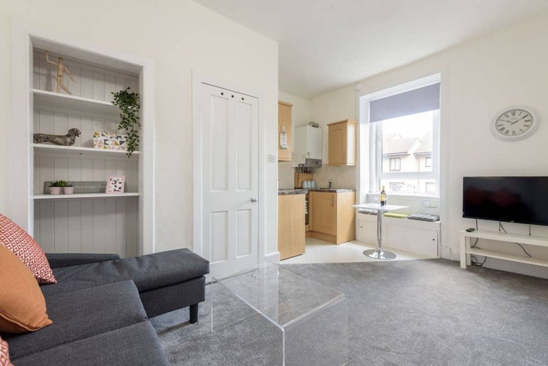 Open-plan living room with grey sofa, shelving unit, kitchen area and window view