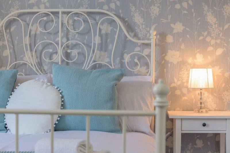 Bedroom with ornate metal bed frame, turquoise pillows, bedside lamp and floral wallpaper