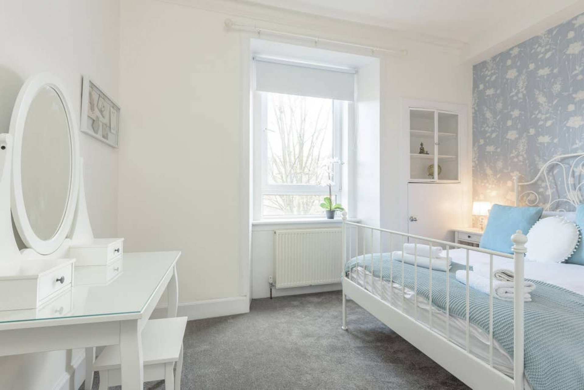 Bright bedroom with single bed, white dressing table, oval mirror, and window with garden view