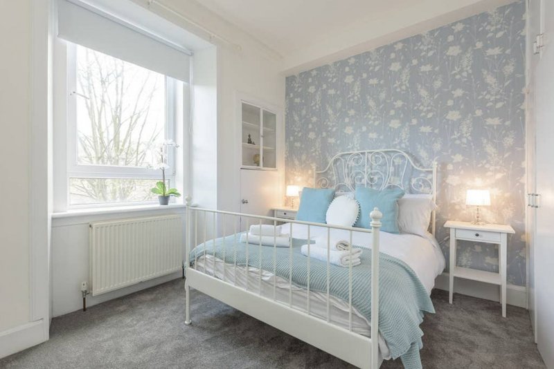 Bright bedroom with white metal bed frame, blue patterned wallpaper, bedside lamps and window