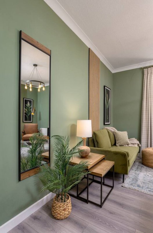 Modern living room with sage green walls, full-length mirror, green sofa, wooden side table, and potted plant