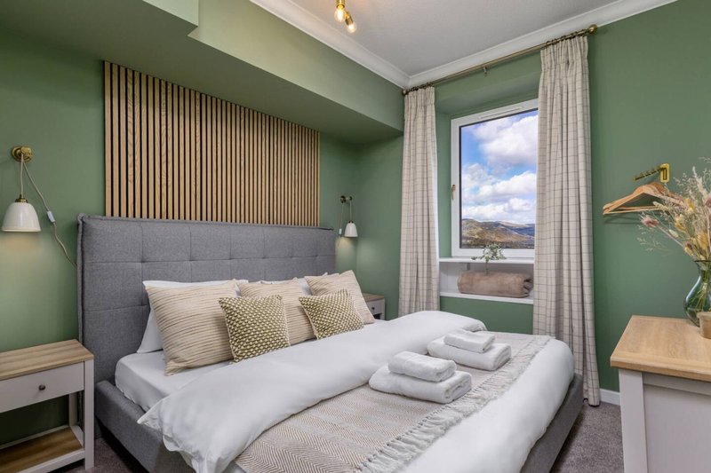Modern bedroom with green walls, grey upholstered bed, white bedding, and window overlooking countryside