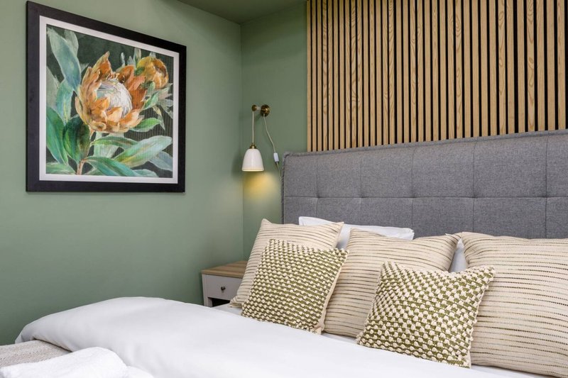Modern bedroom with grey upholstered headboard, patterned cushions, white bedding, and sage green wall