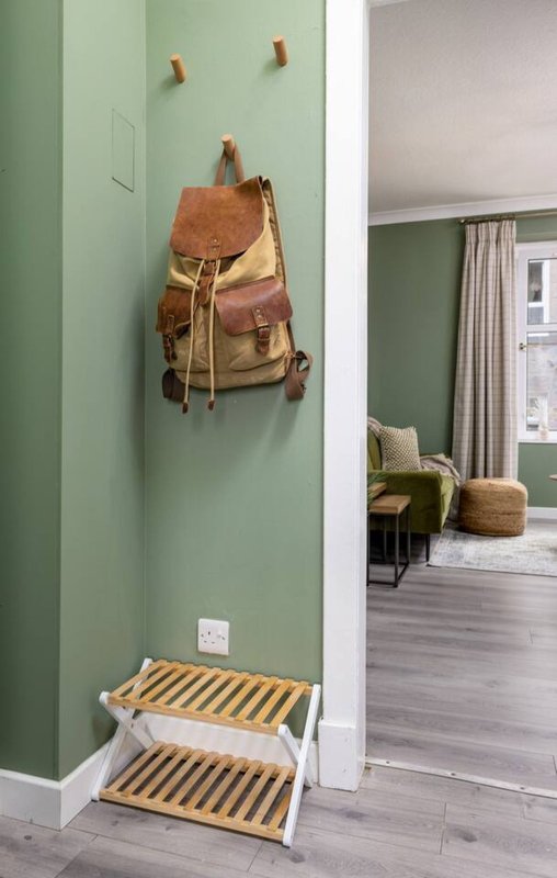 Hallway with sage green walls, leather backpack on hooks, wooden shoe rack, and view into adjacent room